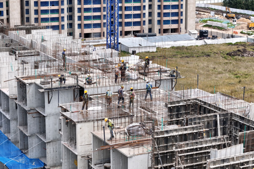 Obreros de la construcción con equipos de seguridad instalan encofrados en la estructura de un edificio de varios pisos con muros de hormigón y armadura vista en una obra urbana.