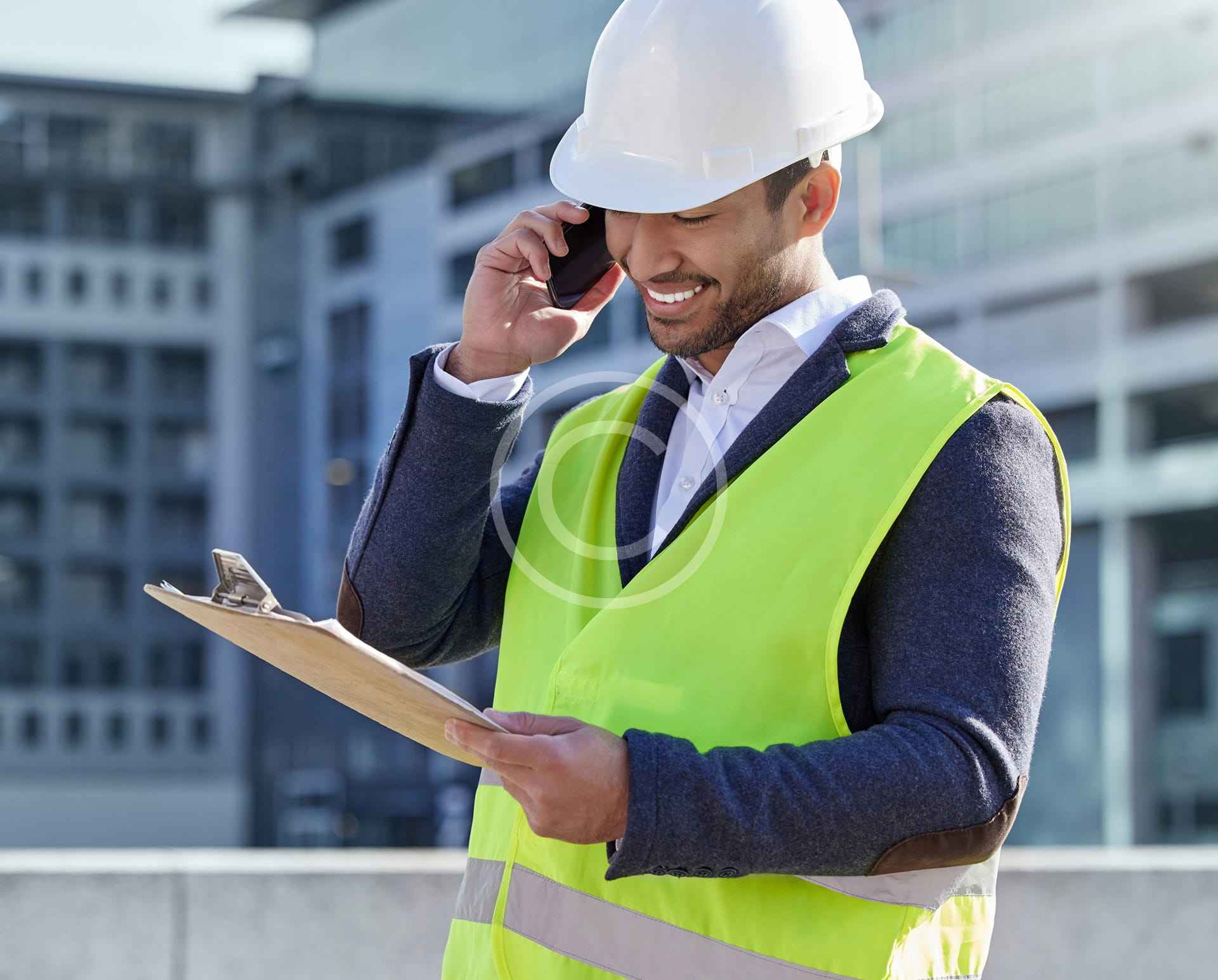 Hombre con casco y chaleco de seguridad amarillo sonríe mientras habla por teléfono y sostiene un portapapeles al aire libre en una obra de construcción, listo para ayudar a entender los códigos de construcción para su guía de propietarios.