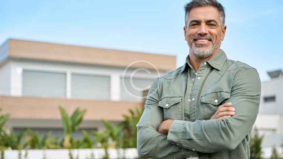 Un hombre de mediana edad con el pelo y la barba grises se encuentra en el exterior de una casa moderna, sonriendo con los brazos cruzados, claramente orgulloso de la reciente renovación de su casa. El fondo muestra vegetación y un cielo azul despejado.