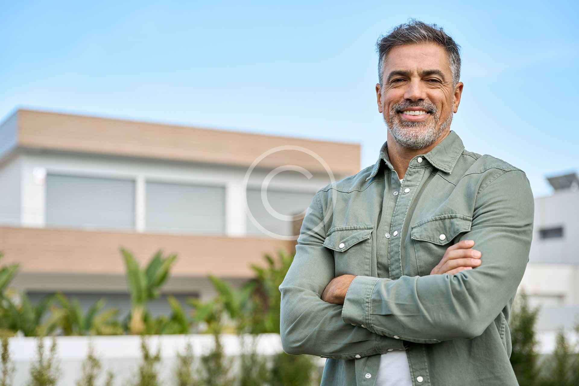 Un hombre de mediana edad con el pelo y la barba grises se encuentra en el exterior de una casa moderna, sonriendo con los brazos cruzados, claramente orgulloso de la reciente renovación de su casa. El fondo muestra vegetación y un cielo azul despejado.