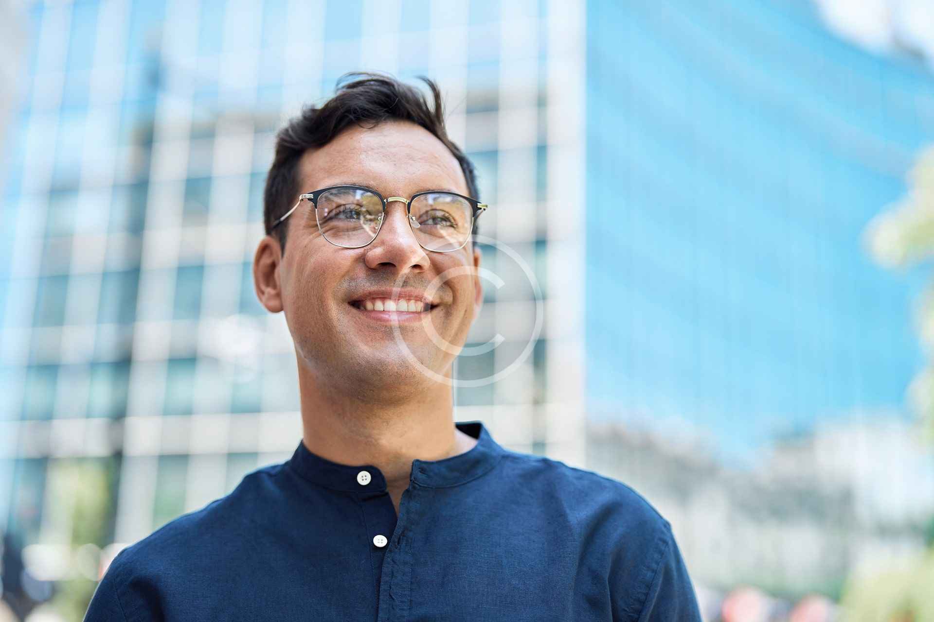 Un hombre con gafas y camisa azul oscuro sonríe de pie frente a un moderno edificio de cristal, reflejo de la evolución del diseño de viviendas y de la construcción moderna.