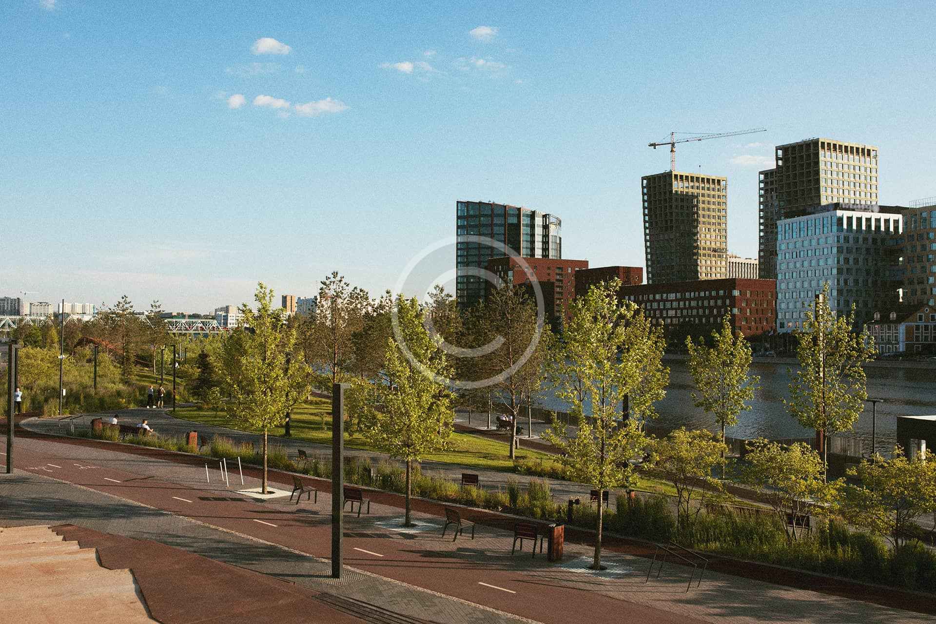 Paisaje urbano con modernos rascacielos de construcción sostenible, un río y un parque con árboles y senderos bajo un cielo azul despejado.