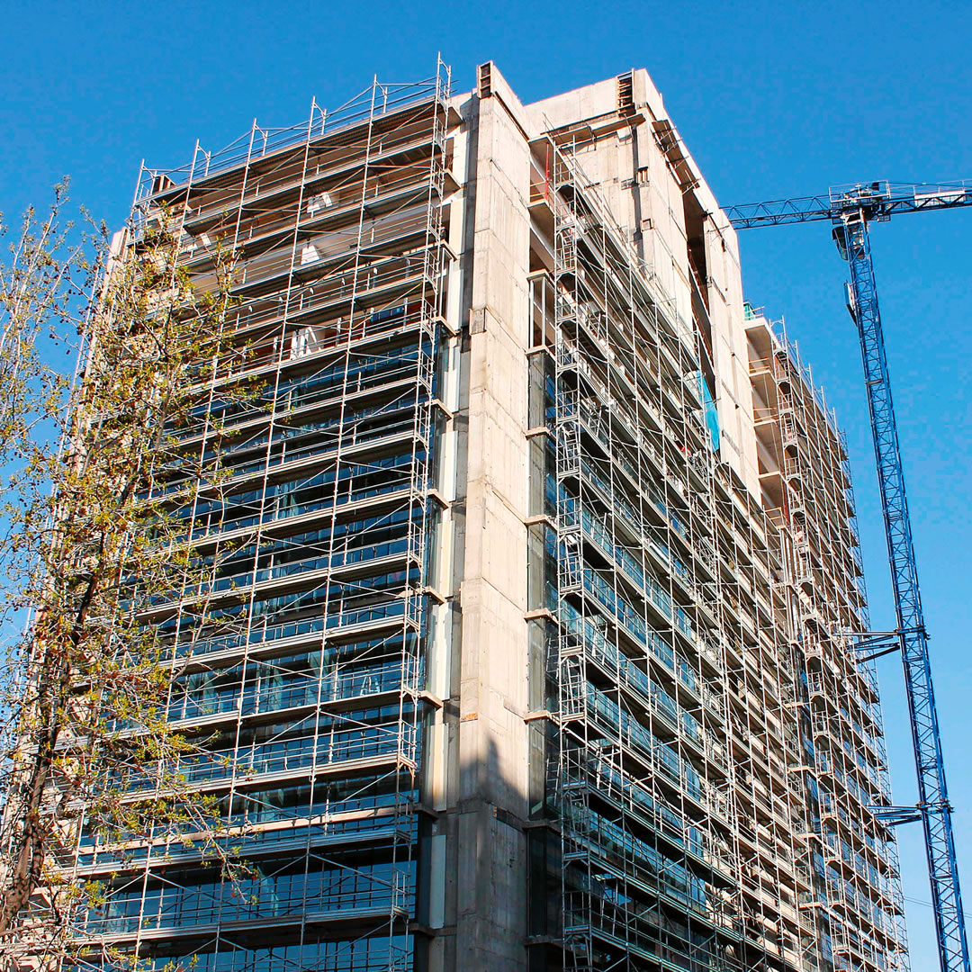 Un edificio alto en construcción, rodeado de andamios, con una grúa azul cerca contra un cielo azul despejado.