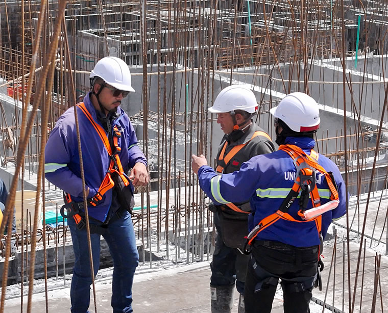 Tres trabajadores de la construcción con casco y equipo de seguridad conversan entre barras de acero en una obra.