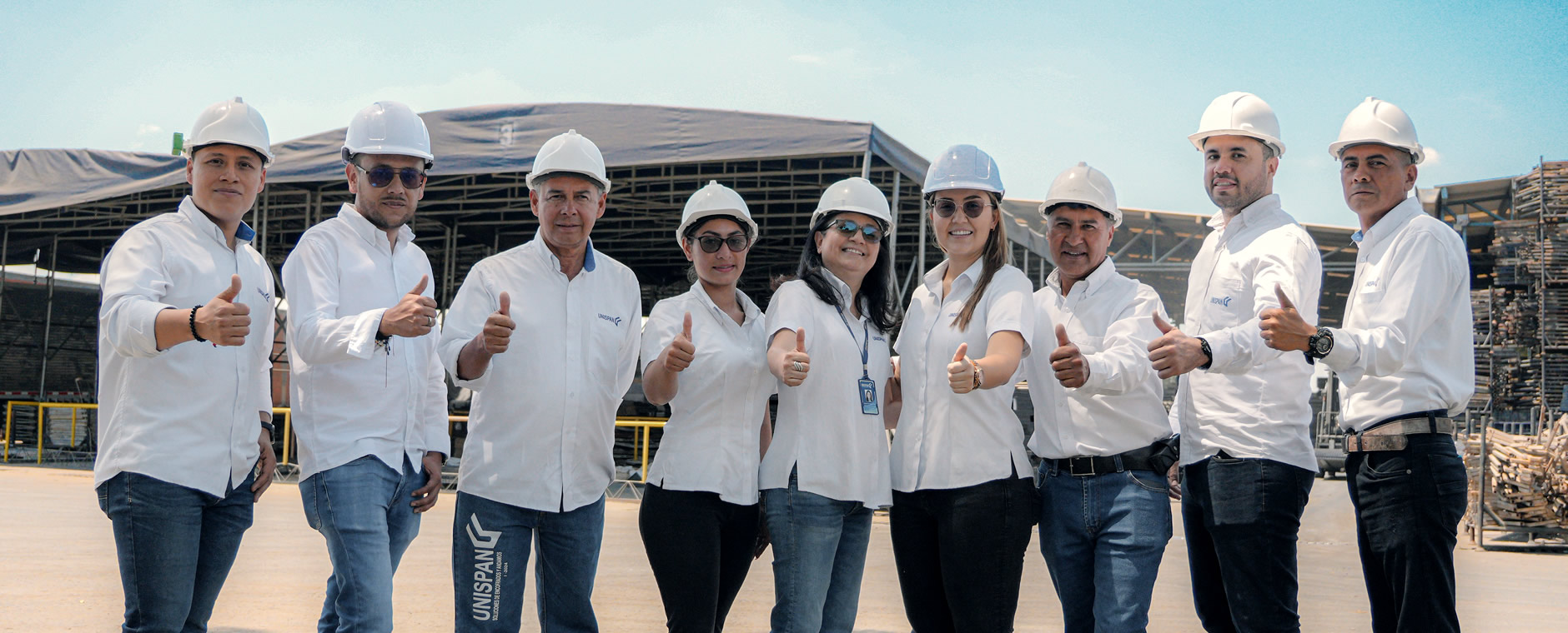 Nueve personas vestidas con camisas blancas, vaqueros y cascos blancos permanecen al aire libre, sonriendo y levantando el pulgar frente a edificios industriales.