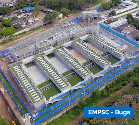 Vista aérea de una instalación grande y moderna con tejados verdes, rodeada de vallas azules y bordeada de árboles. Texto superpuesto: "EMPSC - Buga.