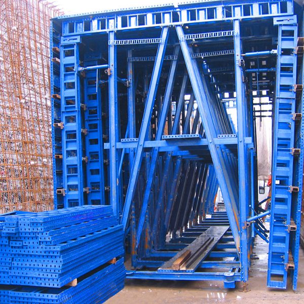 Montones de encofrados y vigas de construcción de metal azul organizados en una obra, con una pila de barras de refuerzo de acero visible a la izquierda.