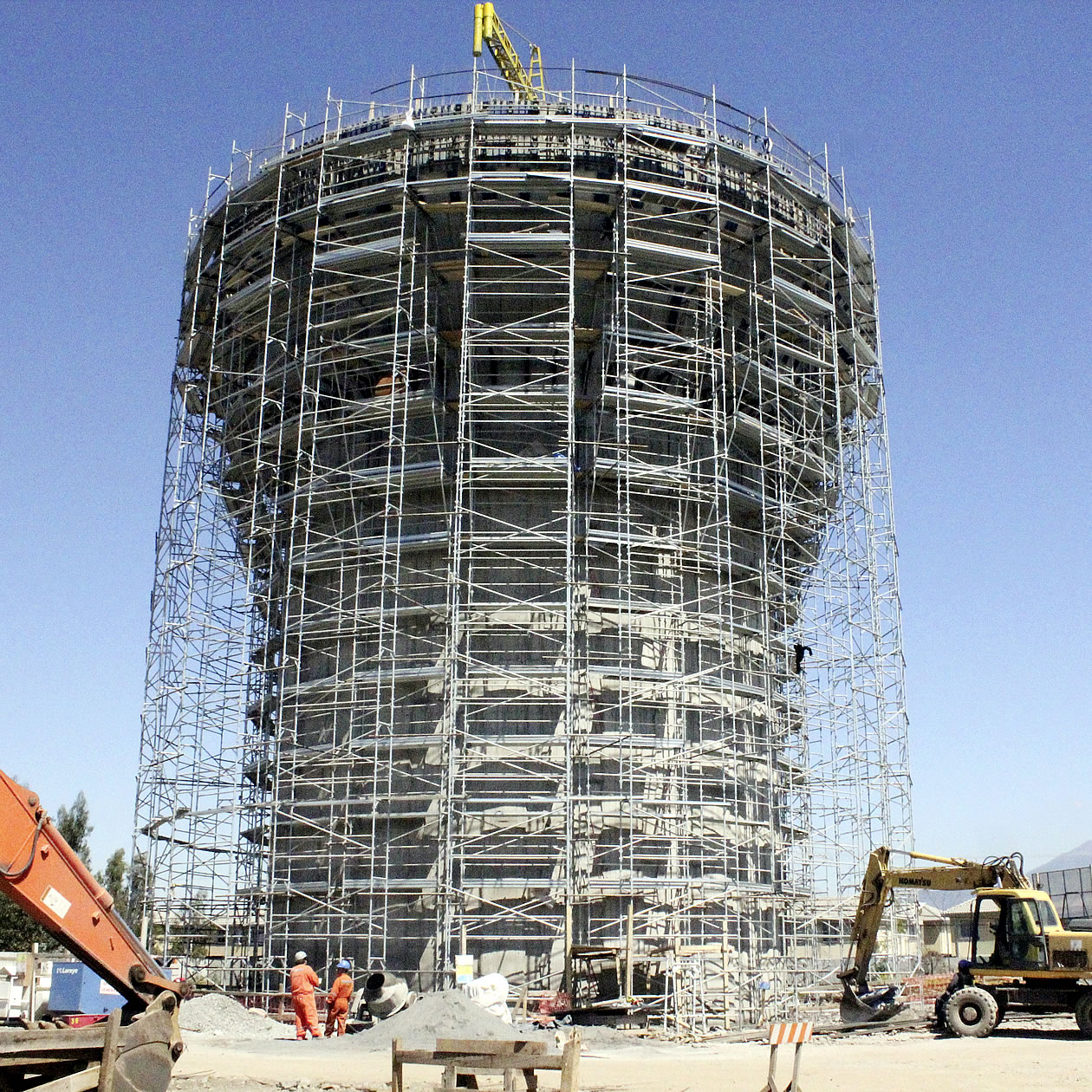 Una gran estructura de hormigón rodeada de andamios en construcción, con trabajadores, equipos de construcción y un cielo azul despejado de fondo.