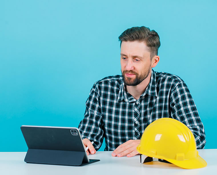 Hombre con camisa de cuadros usando una tableta en un escritorio con un casco de construcción amarillo colocado a su lado, sobre un fondo azul.