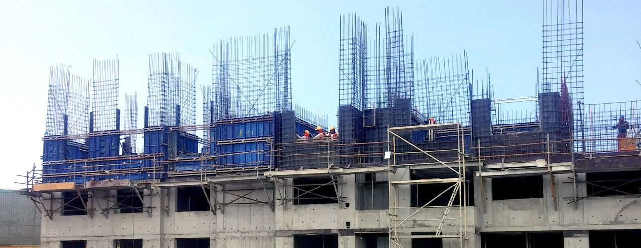 Un edificio de hormigón de varios pisos en construcción con barras de refuerzo a la vista, andamios y varios trabajadores en la obra.