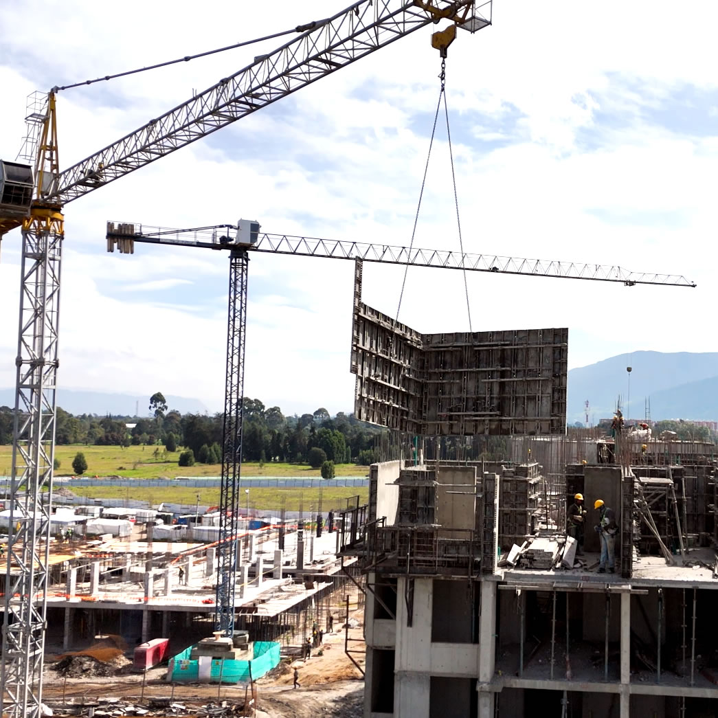 Una obra de construcción con grúas que elevan un gran panel de hormigón sobre la estructura de un edificio de varios pisos, con trabajadores visibles y más obras de construcción al fondo.