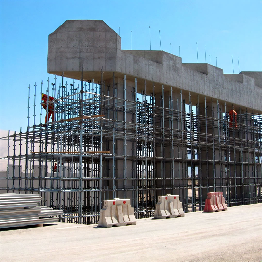 Obra de construcción con obreros en andamios montando una gran estructura de hormigón, con barreras metálicas y materiales de construcción en primer plano.