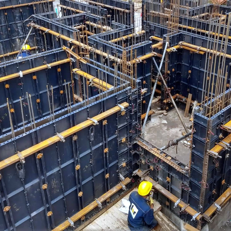 Dos obreros de la construcción con cascos amarillos montan encofrados de acero y madera contrachapada para muros de hormigón armado en una obra.