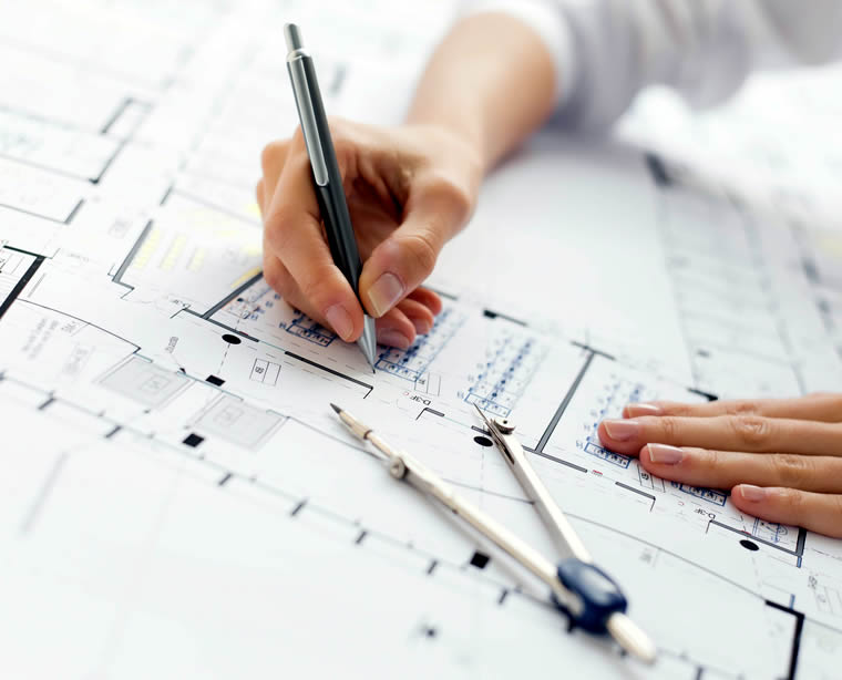 Persona que escribe sobre planos arquitectónicos con un bolígrafo, con una brújula colocada sobre los planos.