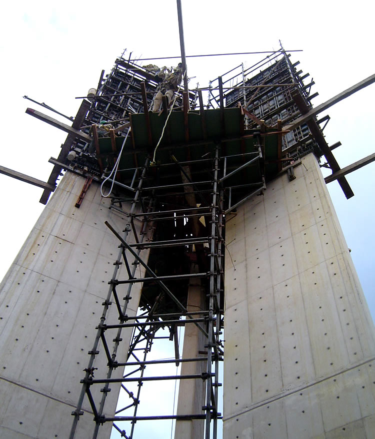 Pilares de hormigón con andamios metálicos y encofrados en construcción, vistos desde abajo contra un cielo nublado.