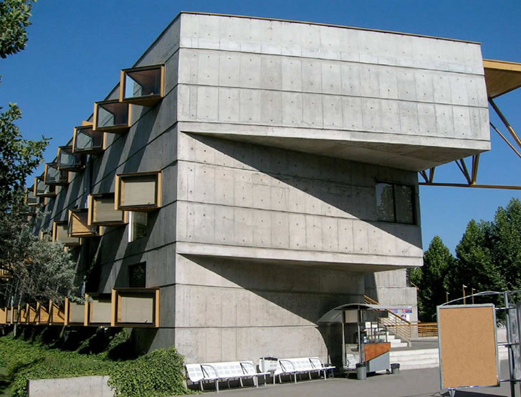Un moderno edificio de hormigón con formas geométricas y ventanas en forma de caja que sobresalen, con una pasarela cubierta y bancos en el exterior. Al fondo se ven árboles y un cielo despejado.