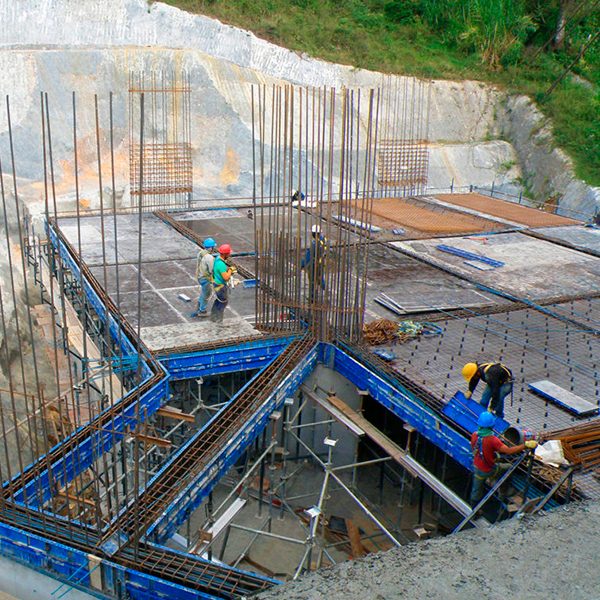 Trabajadores de la construcción montando barras de refuerzo y encofrados en una obra con cimientos de hormigón y andamios, rodeados de tierra y vegetación.