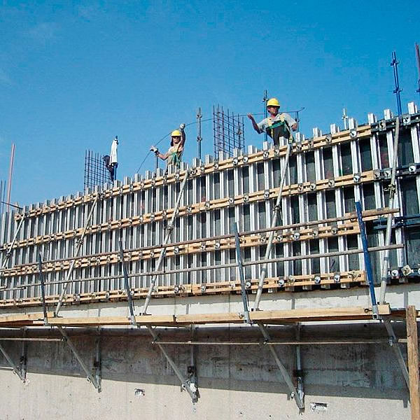Dos obreros de la construcción con equipo de seguridad subidos a un andamio trabajan con encofrados y barras de refuerzo de acero en una estructura de hormigón bajo un cielo azul despejado.