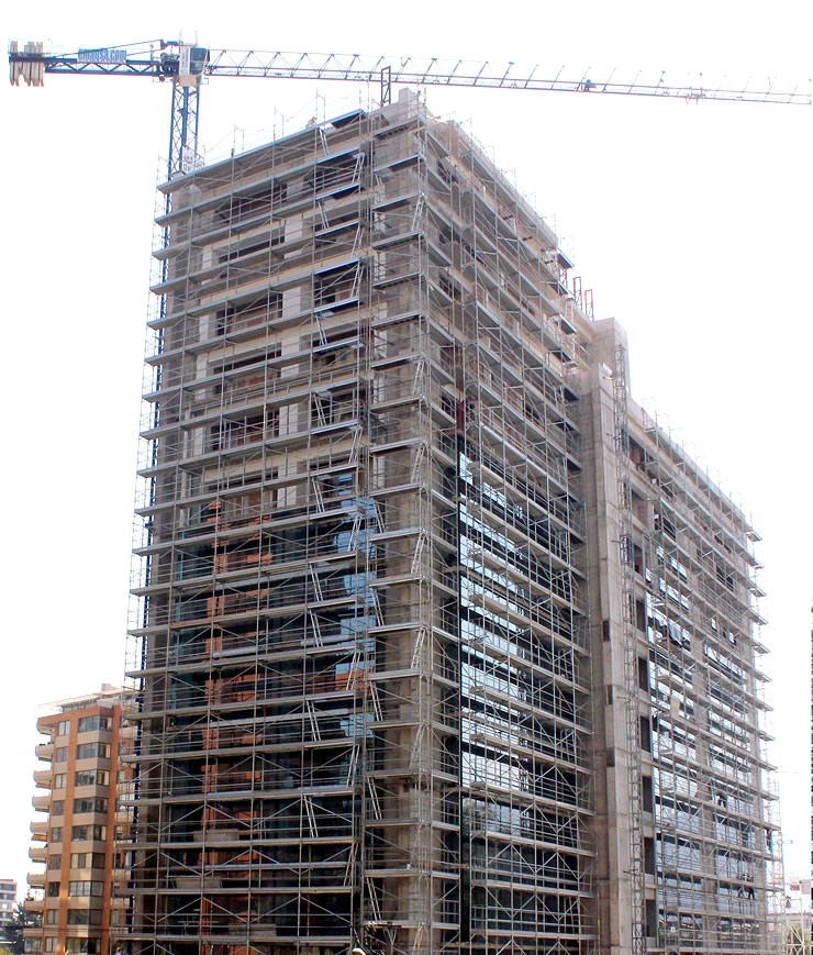 Un rascacielos en construcción, cubierto de andamios, con una grúa torre encima y otros edificios cerca.