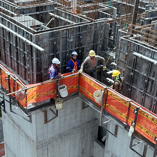 Cuatro trabajadores de la construcción con equipo de seguridad en un rascacielos en construcción, rodeados de encofrados de hormigón y barreras de seguridad naranjas.