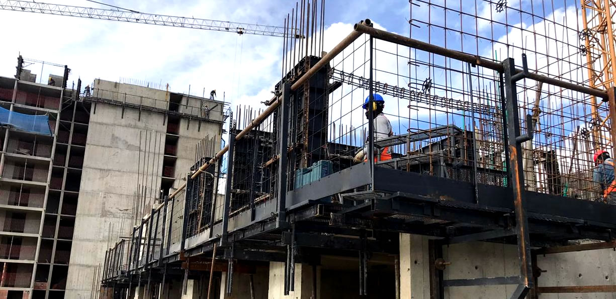 Obreros de la construcción en una obra trabajan en una estructura de hormigón parcialmente terminada con una estructura metálica y grúas de fondo bajo un cielo parcialmente nublado.
