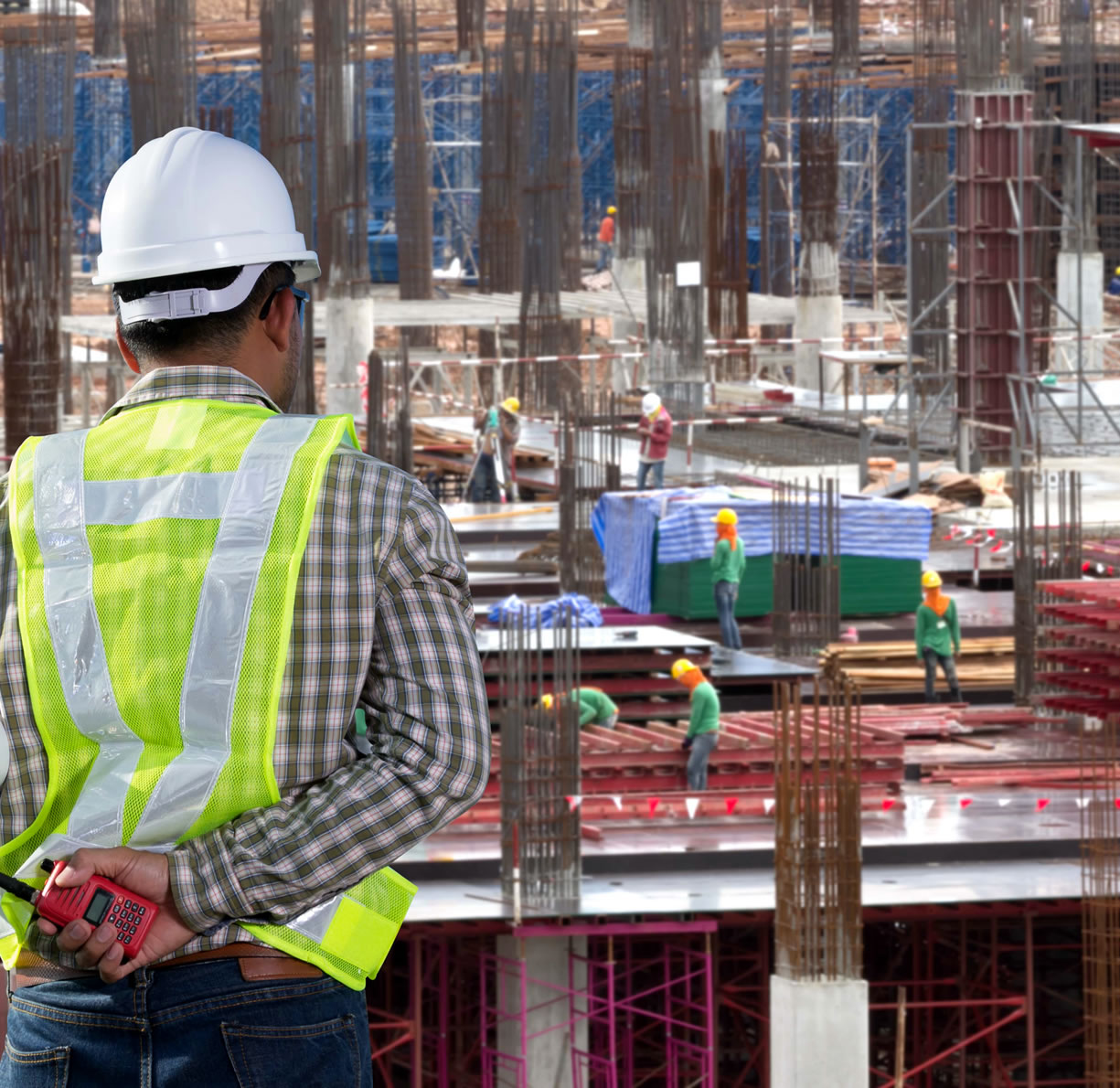 Un trabajador de la construcción con chaleco de seguridad y casco observa una obra con varios obreros y andamios al fondo.