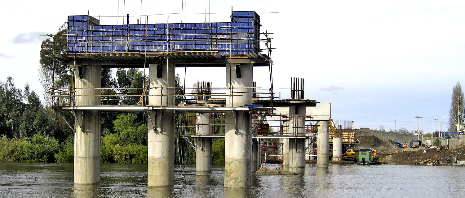 Puente parcialmente construido con pilares de hormigón y andamios sobre un río, con materiales y equipos de construcción visibles en la orilla.
