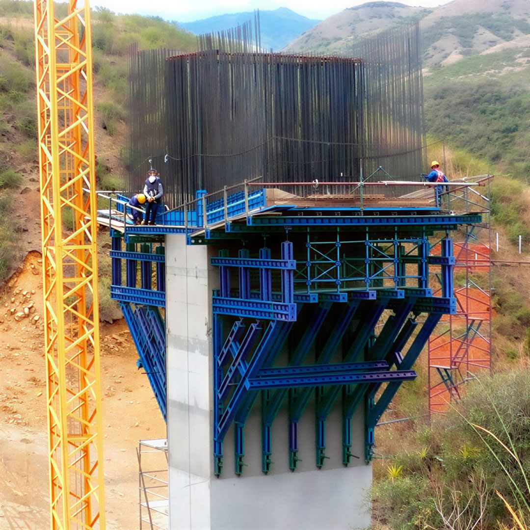 Dos obreros de la construcción de pie sobre un andamio alrededor del pilar de un puente de hormigón armado en construcción en una zona montañosa.