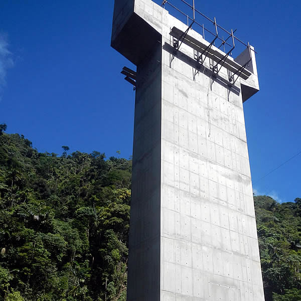 Un alto pilar de hormigón se alza frente a un denso bosque verde bajo un cielo azul despejado.