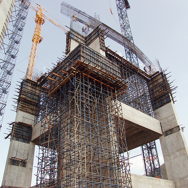 Gran estructura de hormigón en construcción con andamios y grúas, y trabajadores visibles a varias alturas.