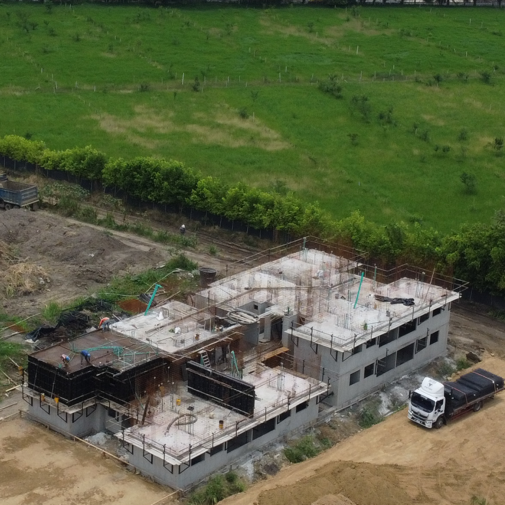 Vista aérea de un edificio en construcción con encofrados y materiales de construcción, rodeado de tierra y campos verdes cubiertos de hierba, con un camión aparcado cerca.