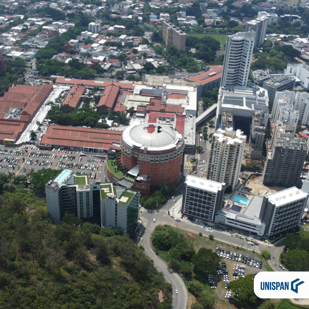 Vista aérea de un paisaje urbano con edificios altos, una estructura central circular, aparcamientos y vegetación circundante. En primer plano se ve una colina. El logotipo de UNISPAN, abajo a la derecha, destaca las soluciones del Sistema Industrializado.