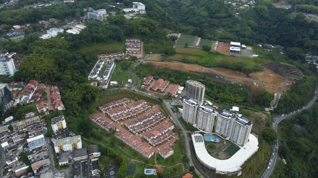 Vista aérea de una zona residencial en Colombia con edificios de apartamentos, casas, zonas verdes, una piscina y carreteras circundantes bordeadas de árboles y terrenos sin urbanizar, con elementos de construcción como el encofrado.