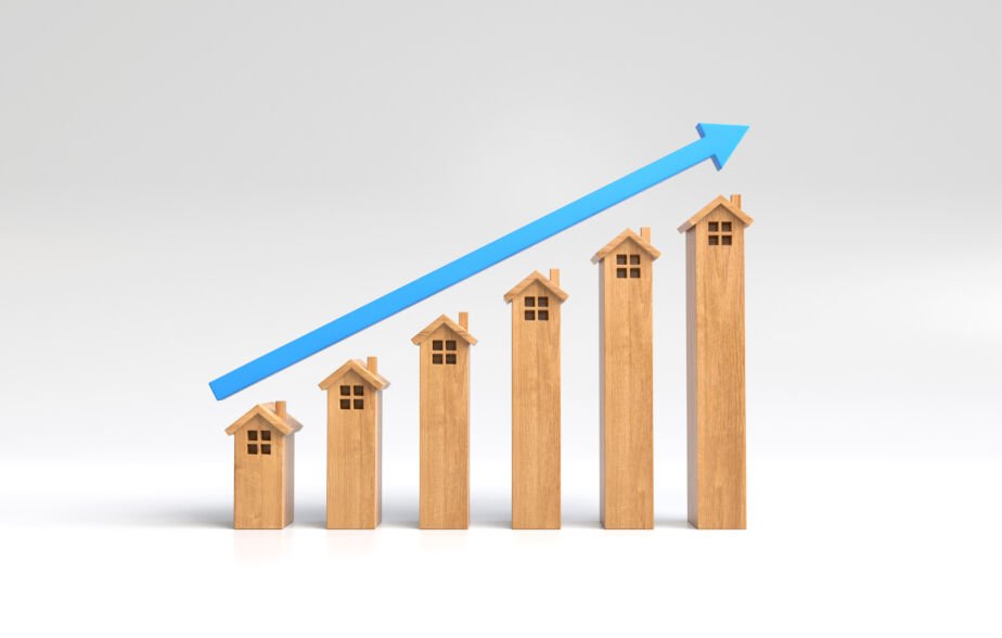 Seis maquetas de casas de madera dispuestas en orden ascendente de altura, con una flecha azul que apunta hacia arriba sobre ellas, lo que simboliza el crecimiento o las tendencias al alza en el sector de la vivienda, el mercado inmobiliario o la tasa hipotecaria.